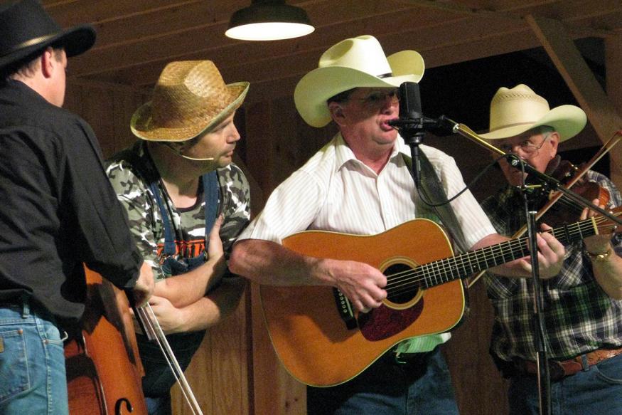 Four men wearing cowboy hats on stage playing a guitar, upright bass, violin, and singing.