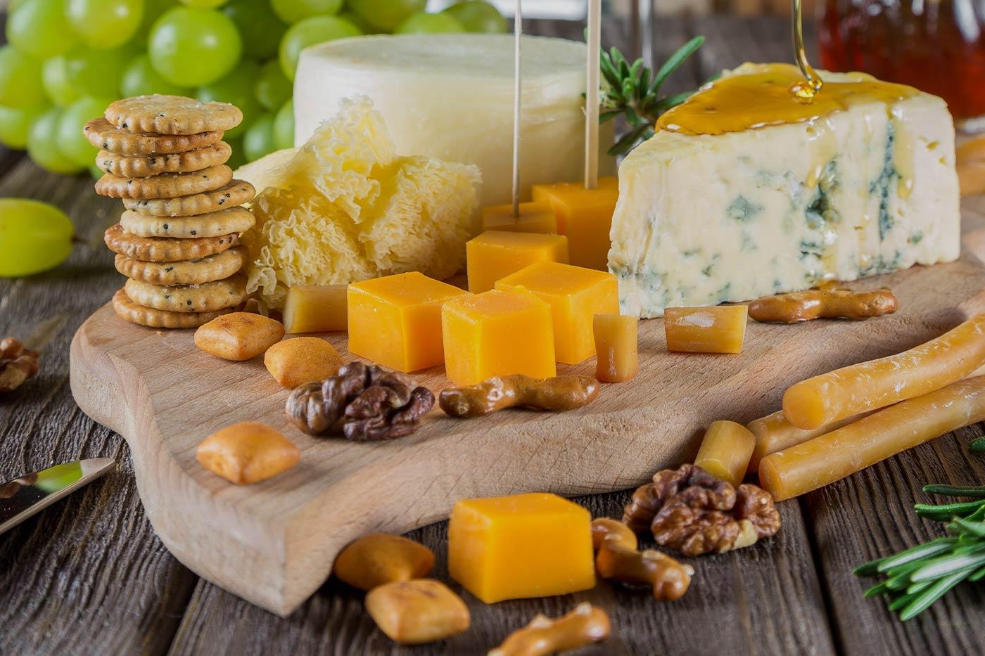 Wooden cutting board covered in walnuts, cheddar cheese, goat cheese, crackers, and other snacks
